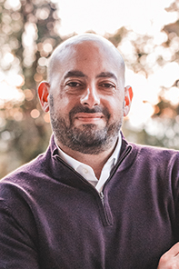 A man with a shaved head and beard, wearing a dark zip-up sweater over a white shirt, stands outdoors with trees in the background.