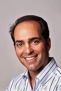 A man with dark hair and a receding hairline is smiling, wearing a striped button-up shirt, in front of a plain light background.