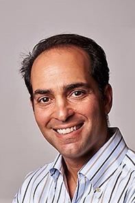 A man with dark hair and a receding hairline is smiling, wearing a striped button-up shirt, in front of a plain light background.