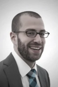 Man with glasses, short hair, and beard wearing a suit, white shirt, and blue checkered tie, smiling at the camera with a plain background.