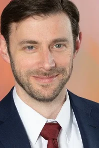 A man with brown hair and a beard is wearing a navy suit, white shirt, and red tie, smiling in front of a blurred orange background.