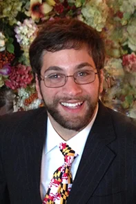 Man with glasses, brown hair, and beard wearing a dark suit, white shirt, and colorful tie, smiling in front of a background of assorted flowers.