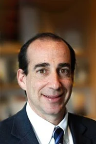A man with short dark hair, wearing a dark suit, white shirt, and striped tie, smiling in front of a blurred indoor background.