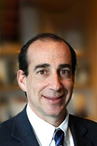A man with short dark hair, wearing a dark suit, white shirt, and striped tie, smiling in front of a blurred indoor background.