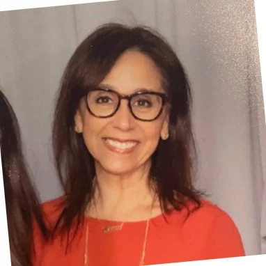 A woman with shoulder-length brown hair and glasses, wearing a red top and necklace, smiles in front of a gray background.