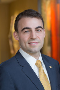 A man in a dark suit and yellow tie poses for a professional headshot, standing indoors with a blurred background.