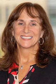 A woman with brown hair, wearing hoop earrings, a necklace, and a patterned blouse, smiles at the camera with a blurred background.