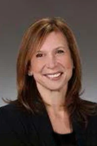 A woman with straight, shoulder-length brown hair is smiling, wearing a dark blazer and a light top, posing in front of a neutral gray background.