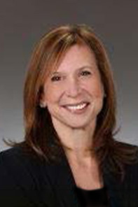 A woman with straight, shoulder-length brown hair is smiling, wearing a dark blazer and a light top, posing in front of a neutral gray background.