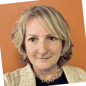 A middle-aged woman with short, wavy blonde hair, wearing a light sweater and a gold necklace, poses in front of an orange background.