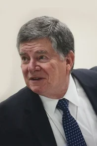 An older man with gray hair wearing a dark suit, white shirt, and patterned navy tie, looking slightly to the side against a plain background.