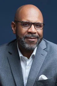 A man with glasses and a beard wearing a grey suit and white shirt poses against a dark blue background.