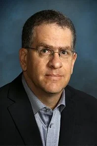 Man with short dark hair, glasses, and a serious expression, wearing a dark blazer over a light blue shirt, posed against a blue-gray background.