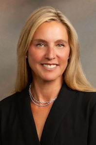 A woman with long blonde hair wearing a black top and a pearl necklace, smiling in front of a neutral background.
