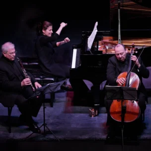 Three musicians perform on stage: a clarinetist, a pianist, and a cellist. The pianist is playing with energy, while the other two read from sheet music and play their instruments.