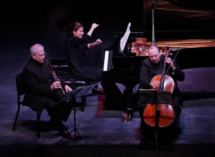 Three musicians perform on stage: a clarinetist, a pianist, and a cellist. The pianist is playing with energy, while the other two read from sheet music and play their instruments.
