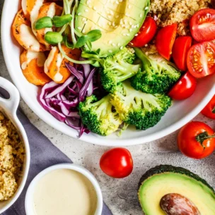 A bowl filled with quinoa, avocado, cherry tomatoes, broccoli, cabbage, and bell peppers sits on a table alongside more quinoa, sliced avocado, broccoli, cabbage, and vine tomatoes.