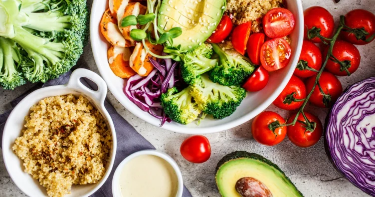 A bowl filled with quinoa, avocado, cherry tomatoes, broccoli, cabbage, and bell peppers sits on a table alongside more quinoa, sliced avocado, broccoli, cabbage, and vine tomatoes.