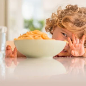 A young child with messy hands sits at a table, peeking from behind a bowl of spaghetti, with a glass of water nearby.