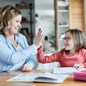 An adult and a child sitting at a table give each other a high five while smiling, with open books and school supplies in front of them.