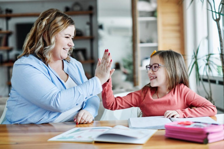 An adult and a child sitting at a table give each other a high five while smiling, with open books and school supplies in front of them.