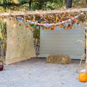 A wooden outdoor canopy decorated with paper garlands, two hanging fabric panels, a bale of hay, a basket of gourds, and three pumpkins, set on a stone patio with trees in the background.