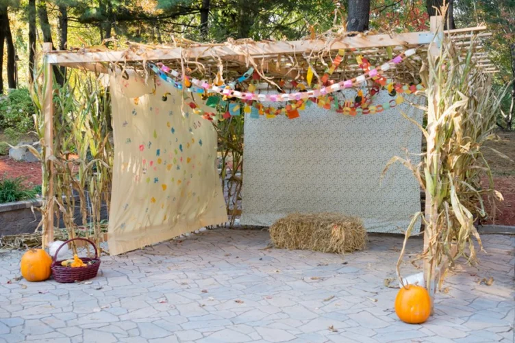 A wooden outdoor canopy decorated with paper garlands, two hanging fabric panels, a bale of hay, a basket of gourds, and three pumpkins, set on a stone patio with trees in the background.