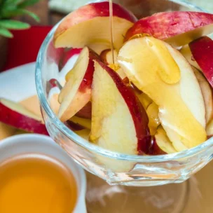 A glass bowl of sliced red apples with honey being drizzled on top, placed on a plate next to a small bowl of honey.