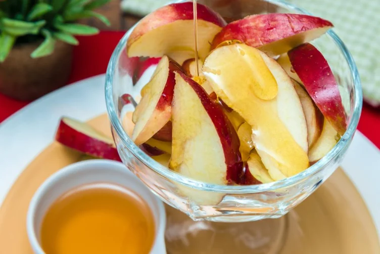 A glass bowl of sliced red apples with honey being drizzled on top, placed on a plate next to a small bowl of honey.
