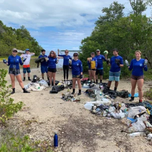 A group of people in blue shirts stand outdoors on a trail, posing with collected trash and debris sorted into piles on the ground. Trees and sky are visible in the background.