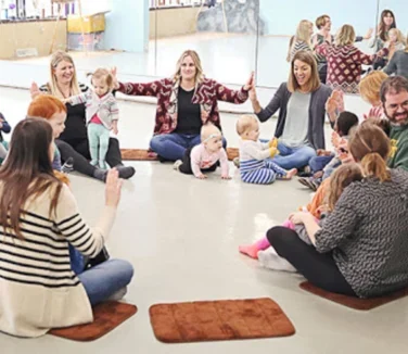 Adults and young children sit in a circle on mats in a brightly lit room, engaging in group activities.