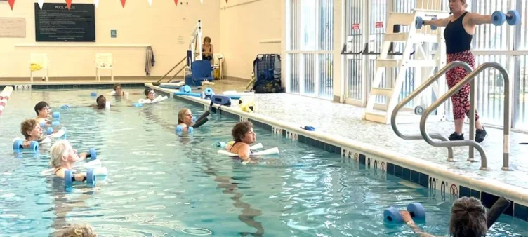 An instructor stands poolside demonstrating water exercise with foam dumbbells to a group of adults participating in a water aerobics class.