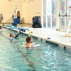 Instructor demonstrates water exercise with foam dumbbells at poolside while class participants follow along in the pool.