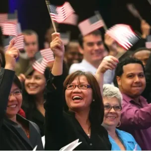 A group of people wave small American flags and smile, appearing to celebrate at an indoor event or ceremony.
