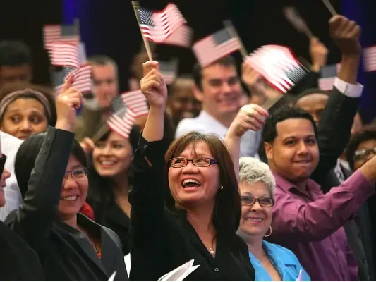 A group of people wave small American flags and smile, appearing to celebrate at an indoor event or ceremony.