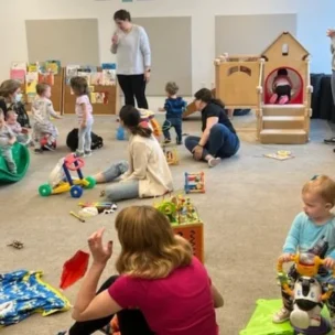 Several adults and young children are engaged in various play activities in a classroom with toys, bookshelves, and a playhouse. Some adults are wearing masks.
