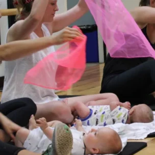 Three women sit on mats indoors, each holding a colored scarf above a baby lying in front of them. The babies are on their backs, and various personal items are nearby.