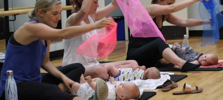 Three women sit on mats indoors, each holding a colored scarf above a baby lying in front of them. The babies are on their backs, and various personal items are nearby.