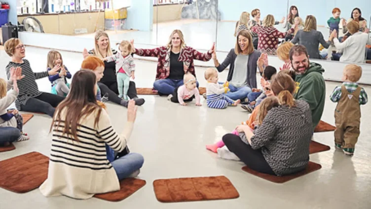 A group of adults and young children sit in a circle on mats in a brightly lit room, participating in a group activity.