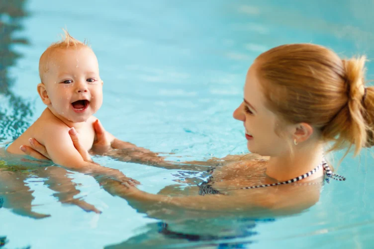 An adult holds a smiling baby in a swimming pool, both partially submerged in the water.