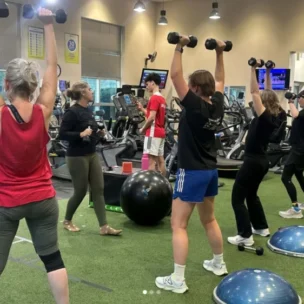 Four people lift dumbbells overhead in a gym while an instructor observes. Exercise equipment, treadmills, and balance trainers are visible in the background.