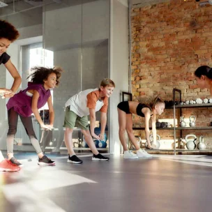 A group of children and an instructor stretch together in a gym with brick walls and shelves holding kettlebells and fitness equipment.