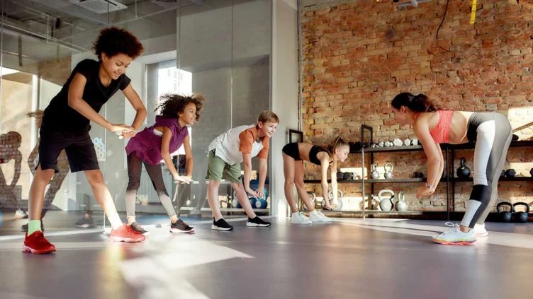 A group of children and an instructor stretch together in a gym with brick walls and shelves holding kettlebells and fitness equipment.