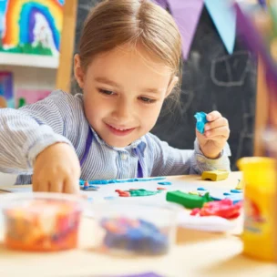 A young child sits at a table and shapes colorful modeling clay, with art supplies and paintings visible in the background.