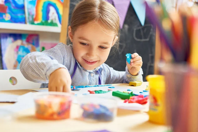 A young child sits at a table and shapes colorful modeling clay, with art supplies and paintings visible in the background.