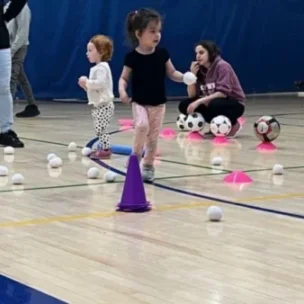 Young children participate in an indoor activity involving plastic balls, cones, and soccer balls on a gym floor, supervised by adults.