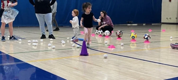 Young children participate in an indoor activity involving plastic balls, cones, and soccer balls on a gym floor, supervised by adults.