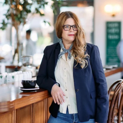 A woman with long hair and glasses, wearing a navy blazer, white shirt, and jeans, stands by a wooden counter in a bright café with a coffee cup nearby.