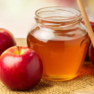 A glass jar of honey with a honey dipper surrounded by three red apples on a wooden surface with burlap fabric.
