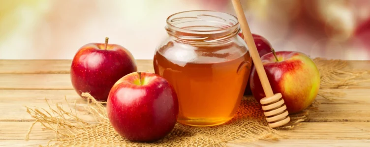 A glass jar of honey with a honey dipper surrounded by three red apples on a wooden surface with burlap fabric.